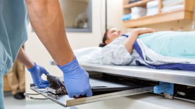 Kvinna ligger på sjukhusbrits och ska genomgå röntgenundersökning. I närbild ett par händer med blåa plasthandskar.