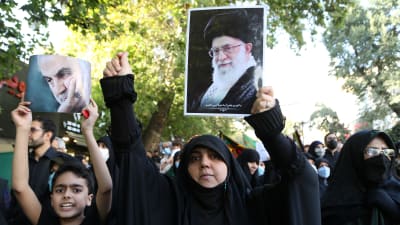 Demonstranter klädda i svart går på en gata och håller i bilder som föreställer iranska ledare och politiker.
