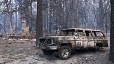 I förgrunden en bil som förstörts i skogsbrand, i bakgrunden ruinerna efter en byggnad.