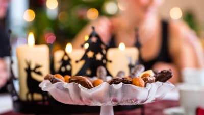 Levande ljus och ett kakfat med julgodis på ett bord framför en julgran.