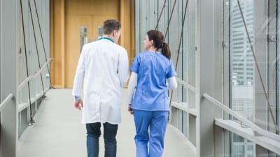 En sjukskötare och en läkare fotograferade bakifrån medan de går i en sjukhuskorridor.
