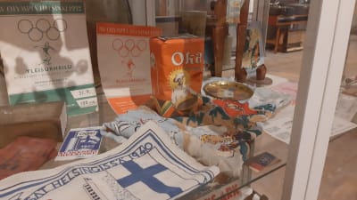 En vitrin i ett museum fylld med saker som hör ihop med OS i Helsingfors 1940 och 1952, såsom programblad, handduk och kaffe.