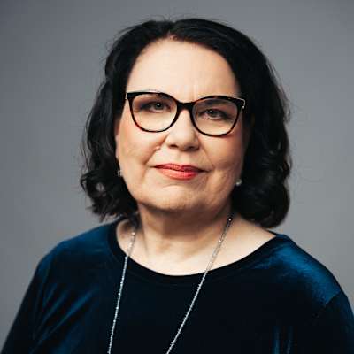 Nainen hymyilee kameralle. Hänellä on yllään tumma samettimekko sekä kaulakoru.