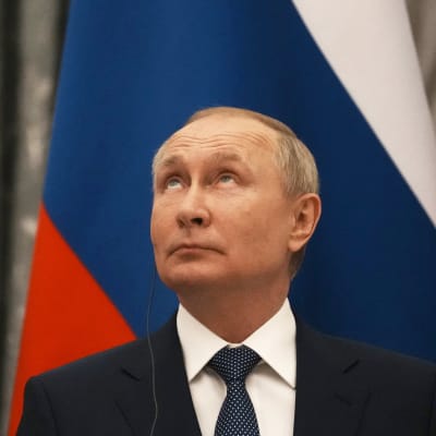 En man i kostym, Rysslands president Vladimir Putin, tittar uppåt framför en rysk flagga.