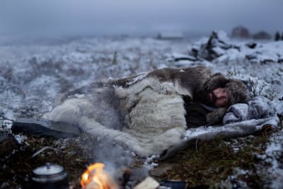 På bilden syns skådespelaren Stig Henrik Hoff ligga insvept i pälsplädar intill en brasa i ett snöigt fjällandskap.