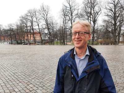 Henrik Saxén, en man med ljusr hår, glasögon och blå rock står en kylig vårdag vid ett torg med nubbstenar. I bakgrunden syns kala träd och en bro.