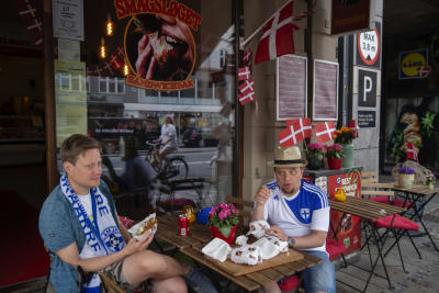 Två män i finska spelskjortor och fotbollshalsdukar sitter på en uteservering prydd med danska flaggor.