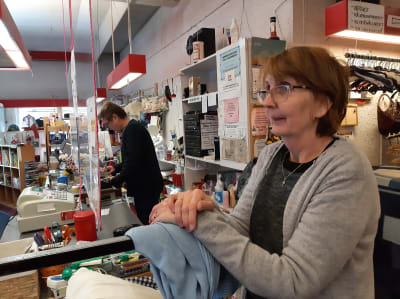 Elina Viljanen, en dam med beige pagefrisyr, glasögon och en grå tröja står i en affärslokal. I bakgrunden står en man vid en kassaapparat.