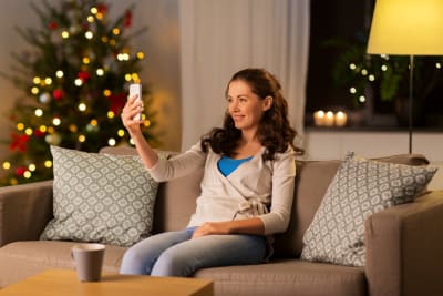 Leende kvinna sitter i en soffa och ser på sin telefon, i bakgrunden en julgran