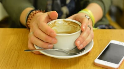Två händer håller i en kaffekopp med mjölkskum. En telefon ligger bredvid på bordet.