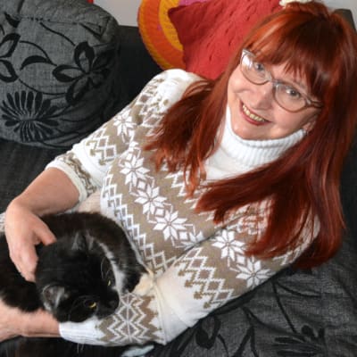 Punatukkainen, silmälasipäinen Saimi Anias kissa sylissään sohvalla.