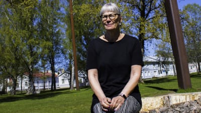 En medelålders kvinna sitter på en stor stubbe på en gräsplan och ler lite. Solen skiner. I Bakgrunden syns gräsmatta, gamla trähus och delar av en klocka samt pelarna som håller upp vajern där klockan hänger.