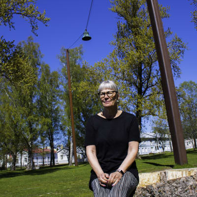 En medelålders kvinna sitter på en stor stubbe på en gräsplan och ler lite. Solen skiner. I Bakgrunden syns gräsmatta, gamla trähus och delar av en klocka samt pelarna som håller upp vajern där klockan hänger.