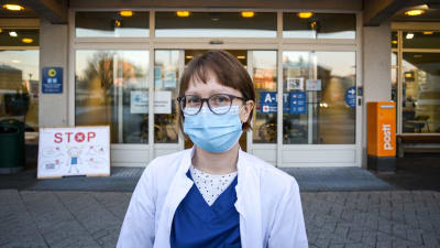 Kvinna i läkarrock och munskydd står framför sjukhusingång och tittar in i kameran.