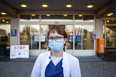 Kvinna i läkarrock och munskydd står framför sjukhusingång och tittar in i kameran.
