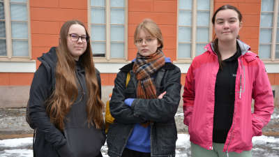 Tre unga personer står utanför en skolbyggnad och tittar rätt in i kameran med ett allvarligt uttryck.