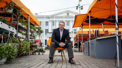 Jan Vapaavuori sitter på en stol på Salutorget, i gången mellan två rader av orangea försäljningsstånd.