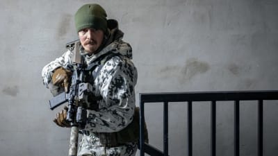 Max Tanner (Jasper Pääkkönen) iklädd en kamouflagedräkt står med ett gevär i handen och ser allvarlig ut.