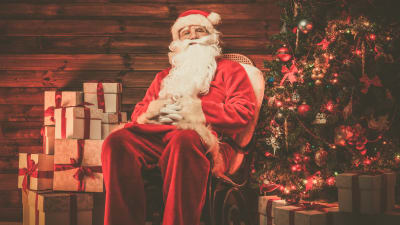 En rödklädd jultomte med långt vitt skägg sitter i en stol invid en julgran och en stor hög julklappar.