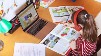 En flicka sitter vid ett skrivbord och skriver i en skolbok. Framför henne en dator.