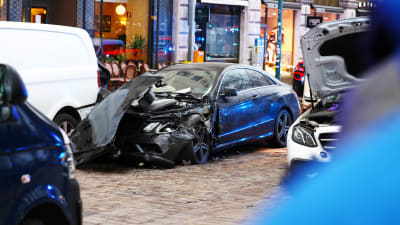 En svart, krockad bil på Norra esplanaden. Fören är bucklig och huven har lossnat och ligger snett uppe på bilen.