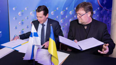 Jan D. Oker-Blom och Oleksandr Menzul sitter vid ett bord och undertecknar ett avtal. På bordet står Finlands och Ukrainas flaggor.