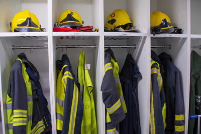 Brandmanskläder hänger galgar i ett omklädningsrum. På hyllan ovanför ligger gula brandmanshjälmar.