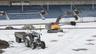 En grävmaskin och en traktor med släp står på Kuppisstadions uppgrävda plan. Massor med snö på marken.
