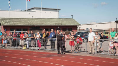 Eri-ikäisiä ihmisiä seisoo kesällä urheilukentän reunalla jotain katsomassa.