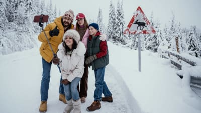 Närbild på en familj som tar ler och tar selfie på en snöig väg, i bakgrunden syns ett trafikmärke som varnar för älg.