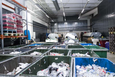En mängd tillplattade plastflaskor i stora sopkärl i ett rum bakom en maskin för att återlämna pantflaskor.