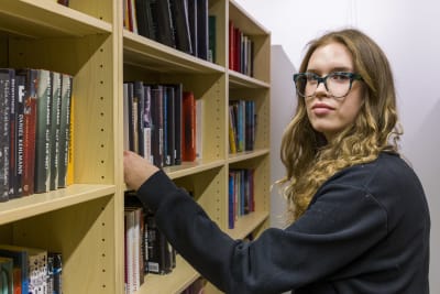 Gymnasieeleven Iris Widbom ser in i kameran vid en bokhylla i Gymnasiet Lärkans bibliotek. 