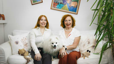 Vivi och Heidi sitter i soffan med hunden som under ett familjeporträtt. Minerna är uppskruvade, stela och påklistrade och tavlor hänger på sned i bakgrunden.