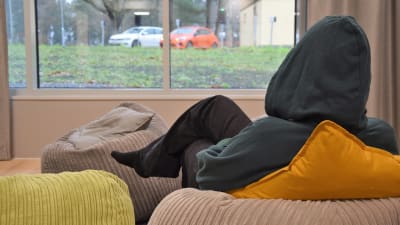 En ung person sitter med en grå munkjacka med huvan dragen över huvudet. Personen har ryggen mot kameran och tittar ut genom ett fönster på en regnig parkeringsplats.