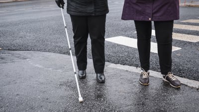 Benen av två personer som står intill ett övergångsställe. Den ena person har i handen en vit käpp som används för personer med synnedsättning.