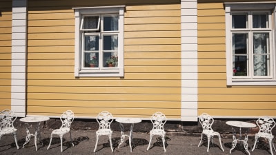 På utsidan av handelshuset. Vita bord och stolar i solskenet framför handelshusets gula vägg.
