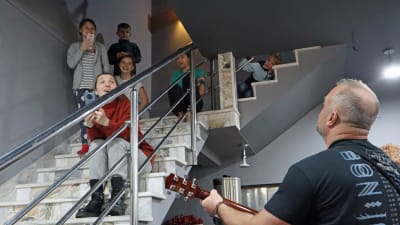 En man med gitarr spelar och sjunger för en grupp barn som sitter på en trappa.