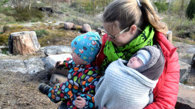 En kvinna sitter utomhus med en sex veckor gamla baby i famnen. Bredvid dem sitter ett två år gammalt barn i en prickig overall.