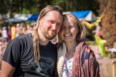 Kaksi ihmistä, mies ja nainen, nojaavat päätään toisiaan vasten ja katsovat kameraan ja hymyilevät Natural High Healing Festivaalilla.
