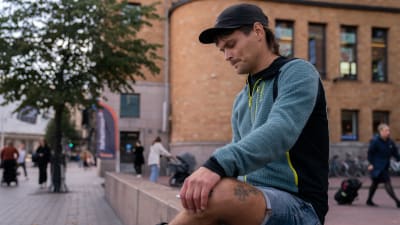 Ung man sitter på bänk i Helsingfors och tittar i sin telefon.