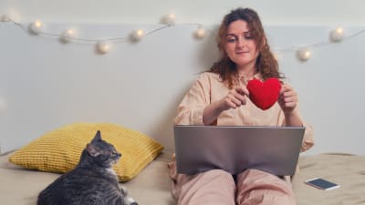 Kvinna sitter på en säng med sin katt och håller i ett tyghjärta. 
