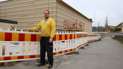 En man i gul tröja står utomhus vid ett bygge. Röd-gult staket runt bygget.
