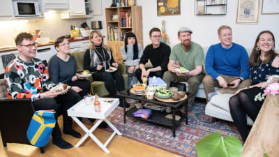 Åtta personer sitter i soffor i ett vardagsrum och ser på tv.