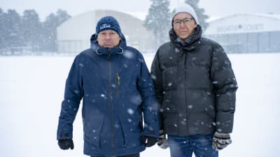 Två män står i snöfall och ser in i kameran.