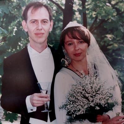 Ett brudpar med glas i hand, tittar båda rakt in i kameran. De är fotograferade utomhus och bruden har en stor bukett färska liljekonvaljer i sin vänstra hand. 