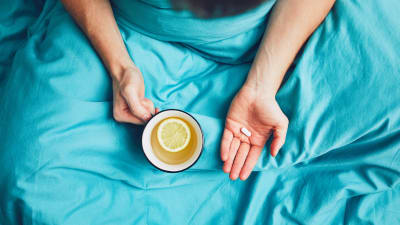 Symbolisk bild för förkylning. En person sitter i sängen med en kopp te och en huvudvärkstablett. 