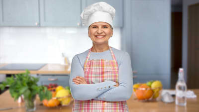 Leende äldre kvinna står i ett kök med kockmössa på huvudet