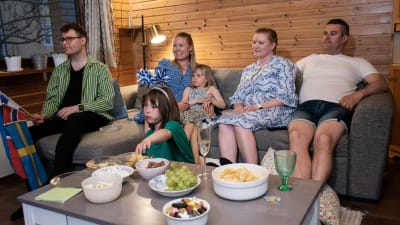 Fyra vuxna och två barn sitter i soffan och kollar på tv. På bordet framför finns olika snacks och godsaker.