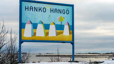 Hangö-skylt vid en strand. På skylten finns fyra ritade badhytter, sol, strand, blå himmel och texten Hanko-Hangö.