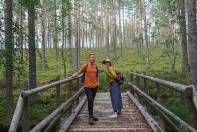 Egenlandin juontajat Hannamari Hoikkala ja Nicke Aldén seisovat puisella sillalla. Taustalla kangasmetsää.